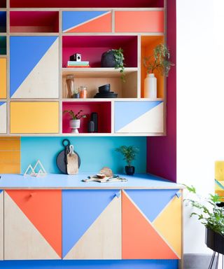 A colorful kitchen cabinet with blue, orange, pink and yellow triangle shapes on it, plants on the shelves, and a white wall next to it