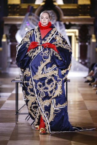 A female model wearing a blue and gold patterned gown with a red shirt under it and gold shoes.