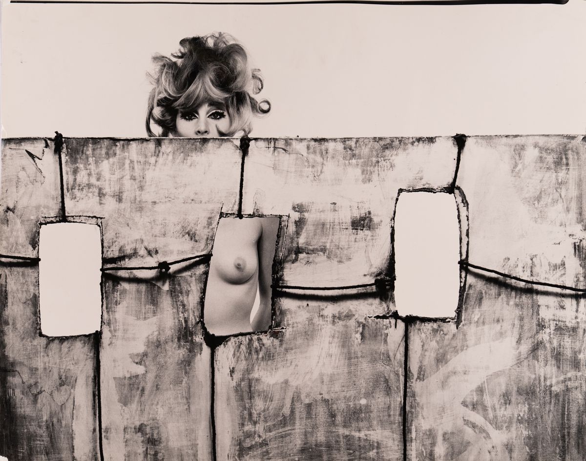 A nude woman crouches on a pale set built with three cut-out window openings, reaching one hand through an opening framed by painted, rope-bound panels.