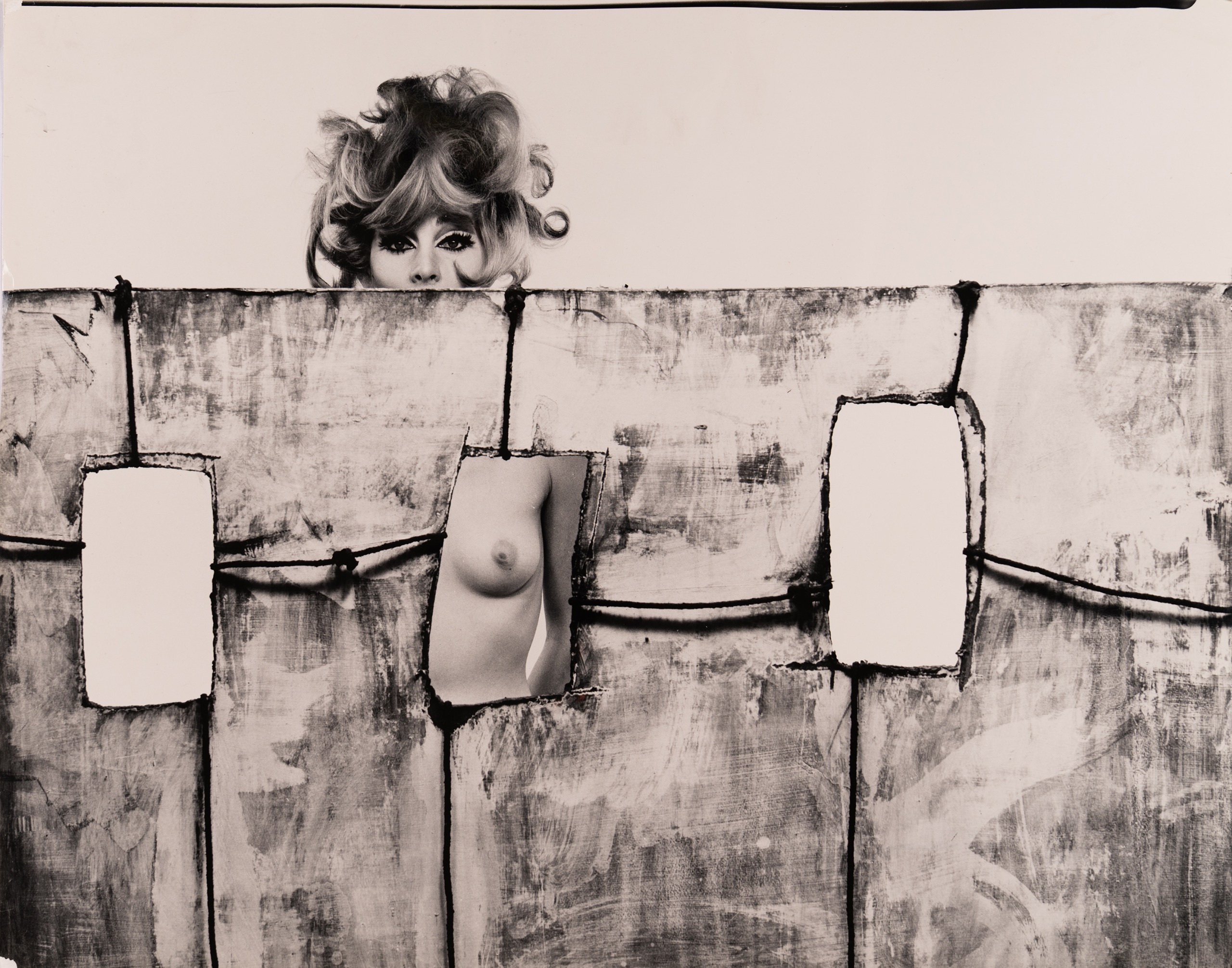 A nude woman crouches on a pale set built with three cut-out window openings, reaching one hand through an opening framed by painted, rope-bound panels.