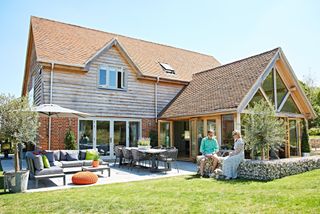 A couple sat outside a red-brick, part oak frame and flint-clad house with outdoor entertaining area