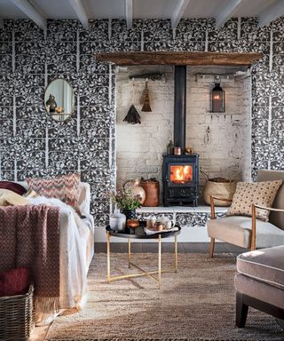 Cozy, country living room with black and white floral wallpapered wall, fireplace with compact wood burning stove, fireplace accessories surrounding the area, oval mirror on wall, dark wooden flooring, two armchairs and sofa facing each other, covered in cushions and blankets