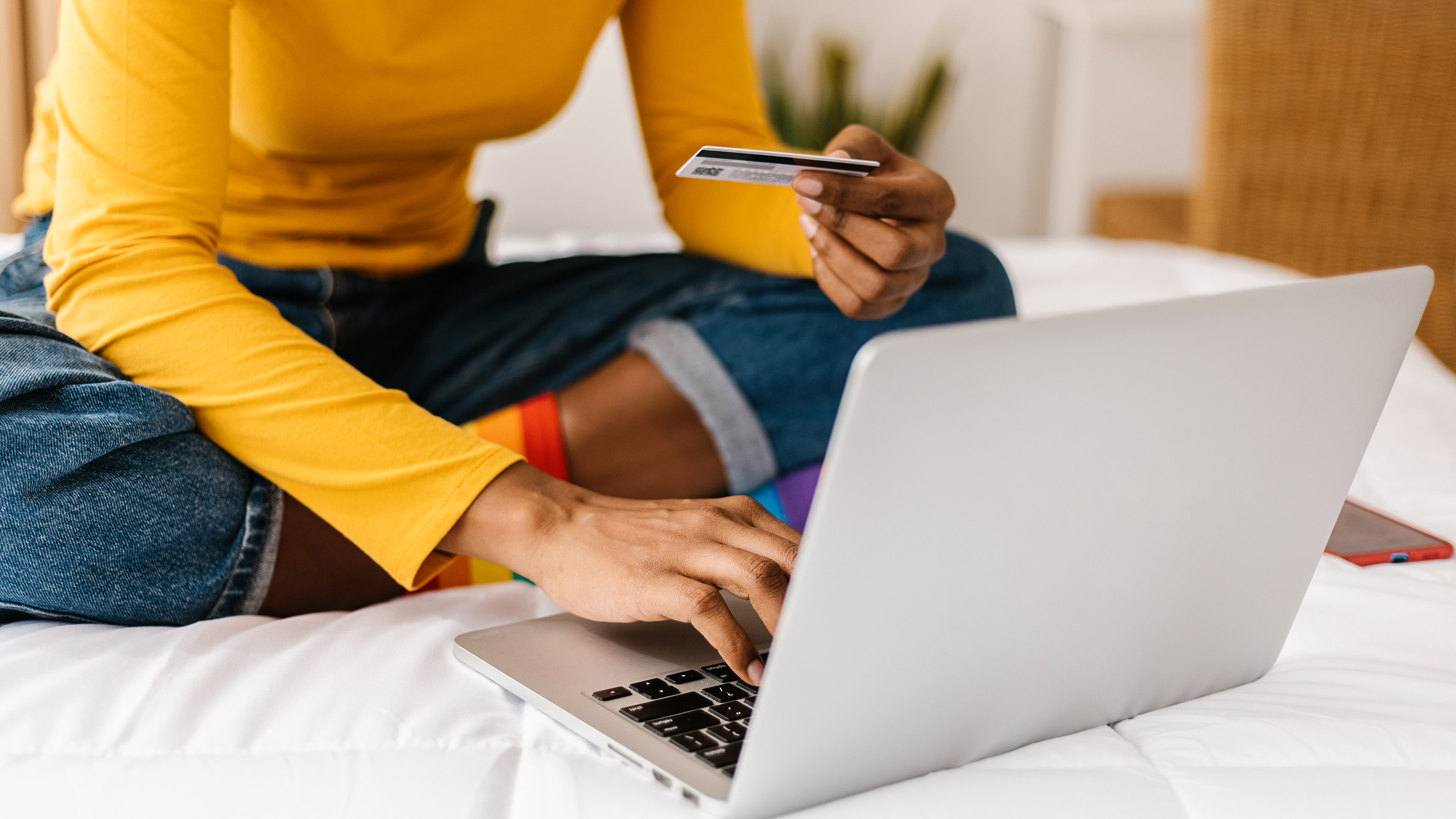 A female figure holding a credit card and typing on a laptop while sat on a bed. Her head is not visible