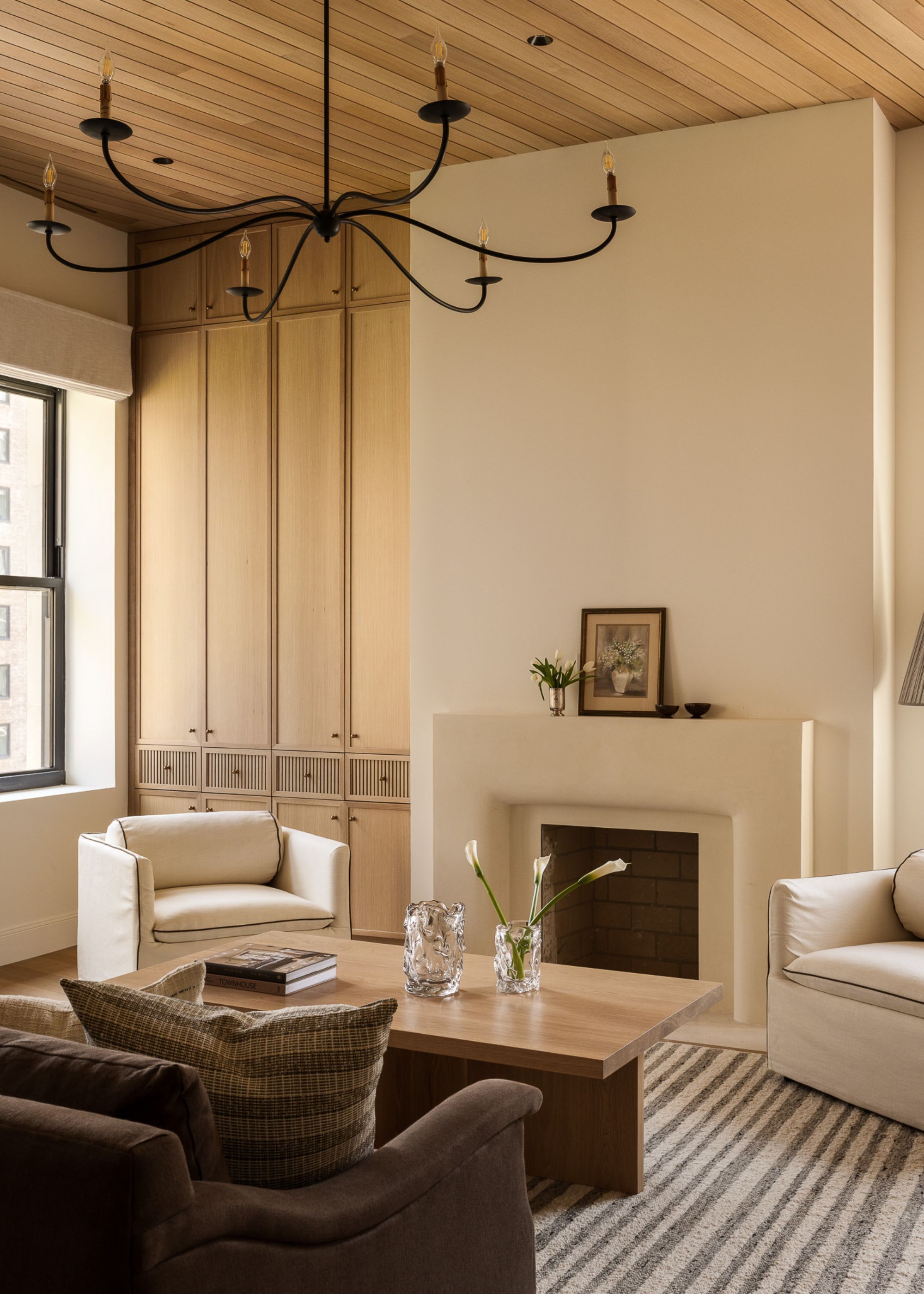 A modern neutral living room with built-in storage, cream walls, wooden paneled ceiling, a slim, modern, black-lined chandelier and a striped rug below the brown sofa and white armchairs with piping