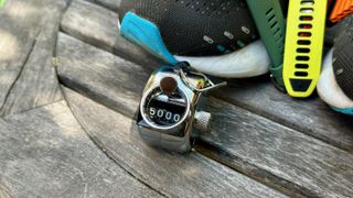 An Ultrahuman Ring Air smart ring sitting on top of a pedometer for step counting showing 5,000 steps. They both sit on a table in front of a shoe and a pile of smartwatches.