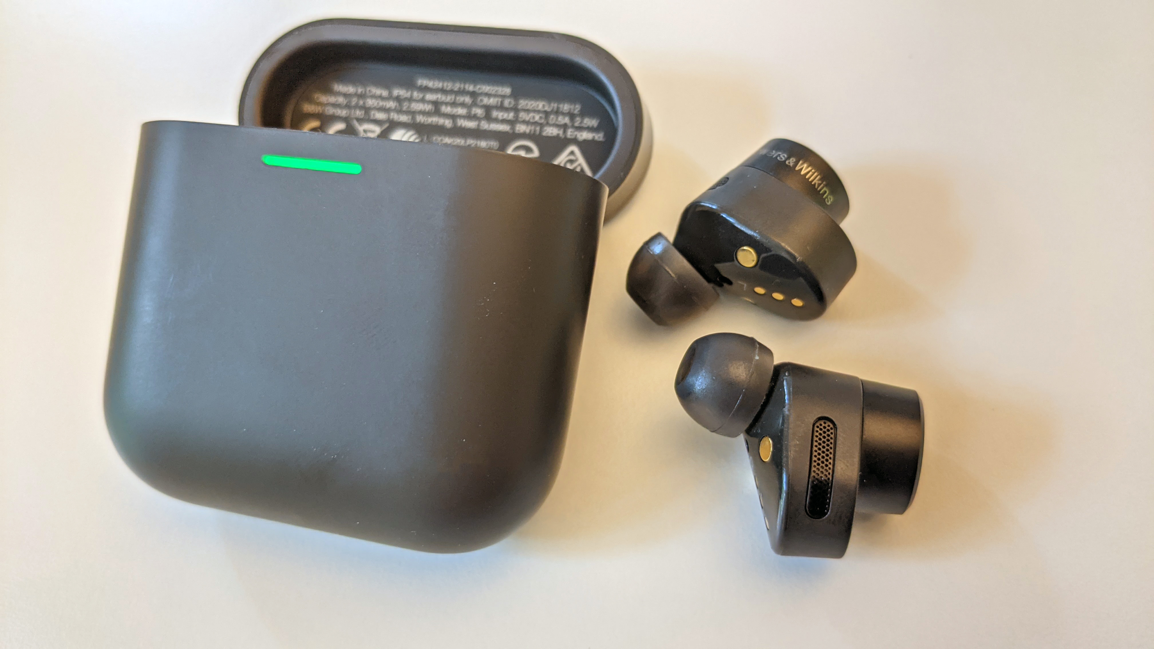Anthrazitfarbene Bowers & Wilkins PI5 liegen neben dem Ladecase auf einem weißen Tisch