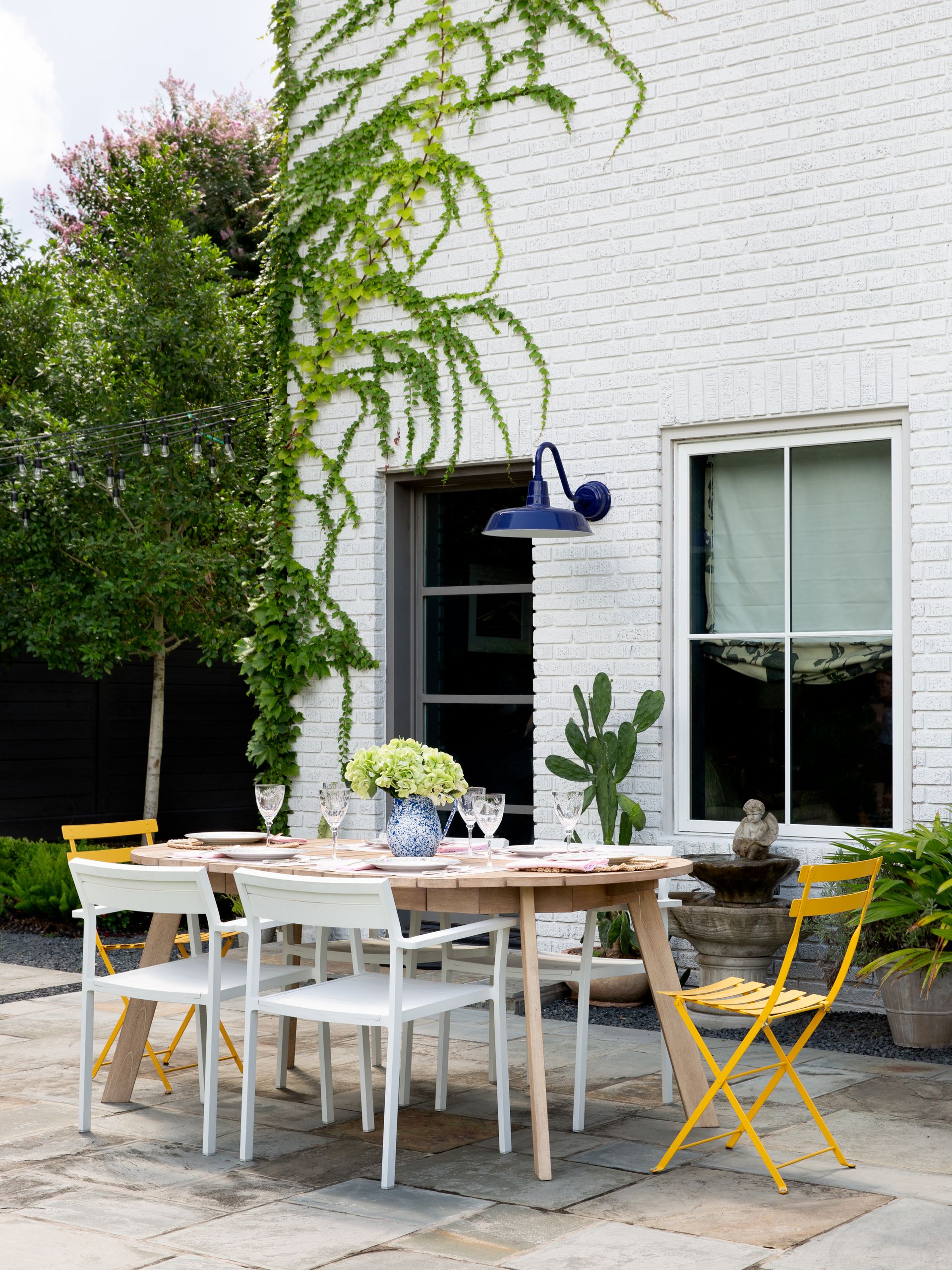 A garden with a wooden table and mismatched seating in white and yellow, and a blue barn light on the white-painted brick facade of the house with climbing foliage