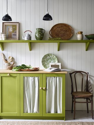 Green painted kitchen cupboards, fabric curtains on cupboard doors, matching green painted shelf decorated with art, ornaments and kitchenware, white paneled wall, black hanging pendants, traditional wooden chair, white flooring with natural rug.