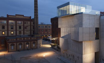 On the right of the image is the exterior of an abstractly designed sandy-coloured building with floor to ceiling glass design on the topmost layer. On the left is two-toned building in 2 shades of brown 