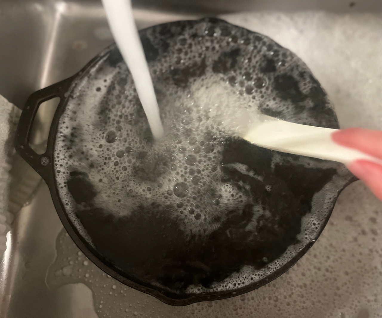 A cast iron pan filling with water and being cleaned with a brush in a stainless steel sink.