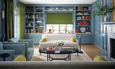 a blue painted living room with custom bookshelves and storage surrounding the window with a white couch, two blue patterned armchairs and a black round coffee table