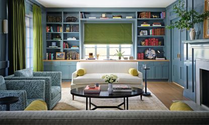 a blue painted living room with custom bookshelves and storage surrounding the window with a white couch, two blue patterned armchairs and a black round coffee table