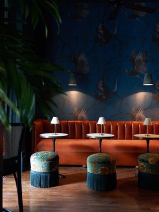 A sumptuously decorated hotel features old-style velvet banquettes and puffs, white Art Deco table lights and round tables, along with birds-filled blue wallpaper.