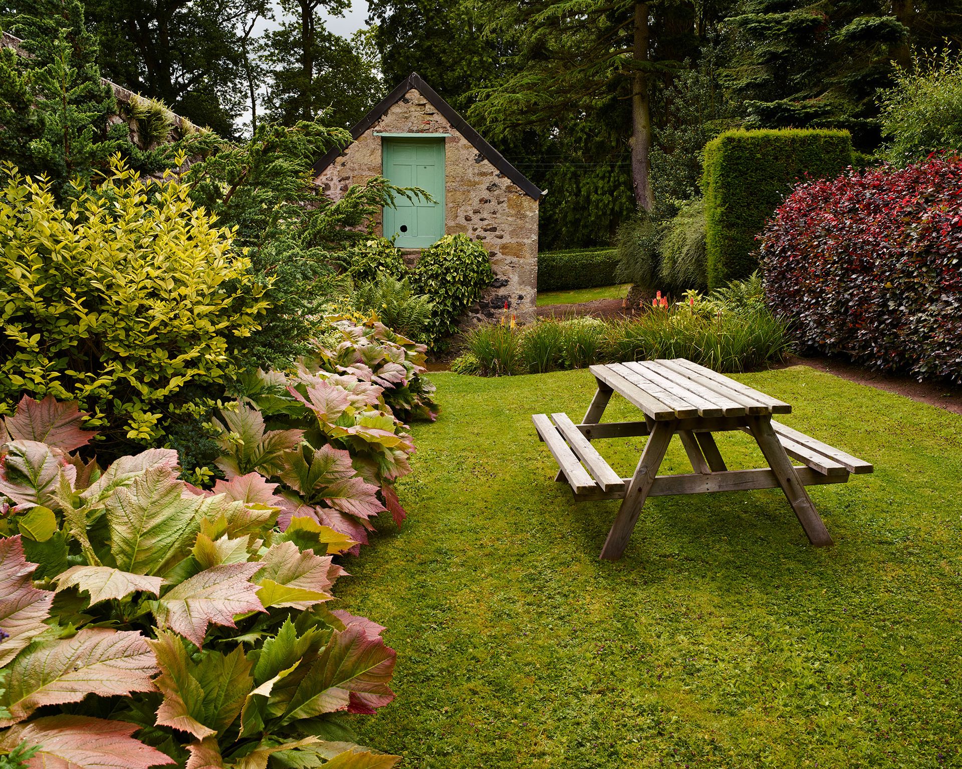 A sloping garden with a wooden dining bench on the lawn in front of a small brick building