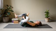 woman on her back on a grey exercise mat performing a hip stretch with a plain wall and plants behind her. 