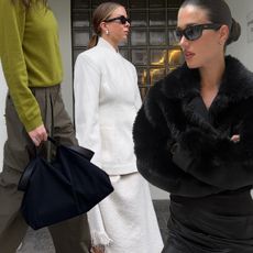 one woman wears a green sweater and pants with a black bag; one woman wears a white blazer and skirt; one woman wears a black fur coat and a black dress.