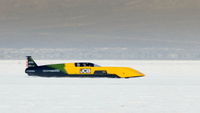 A black, green, and yellow streamlined vehicle on a bright salt flat.