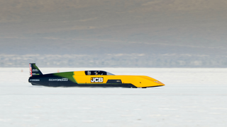 A black, green, and yellow streamlined vehicle on a bright salt flat.