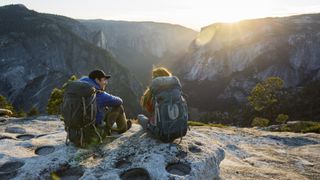 A man and woman sit on a cliff in Yosemite wearing backpacks and looking at the view