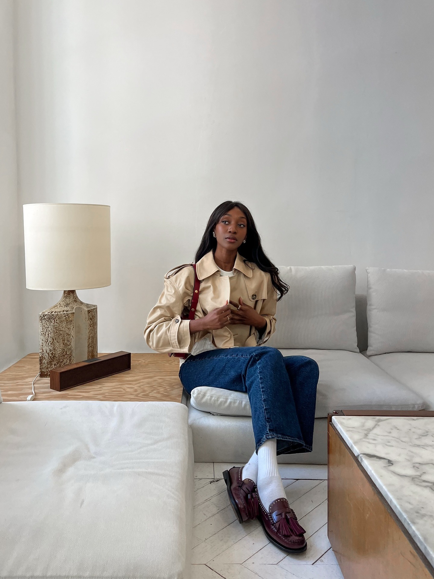 Fashion person Emma wears burgundy loafers with white socks, blue jeans and a cropped trench. she is sat on a sofa in a recent image from her Instagram.