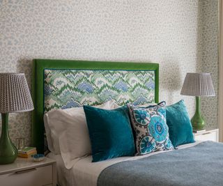 green and blue patterned headboard, blue cushions on bed and pale green floral patterned wallpaper behind bed