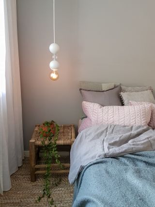 White bedroom with little light, coir flooring, rattan and wood stool/bedside, pastel blue and pink bedding, plant