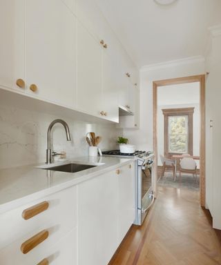 A white kitchen with white cabinets with wooden handles, an integrated sink, a silver oven, wooden flooring, and a wooden door frame looking into the next room