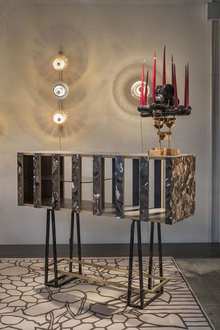 A wooden candelabra stands on metal legs. The wood almost looks like it has an army jacket pattern in neutral tones. On top of it is a candle stick in black and gold, with red candles.