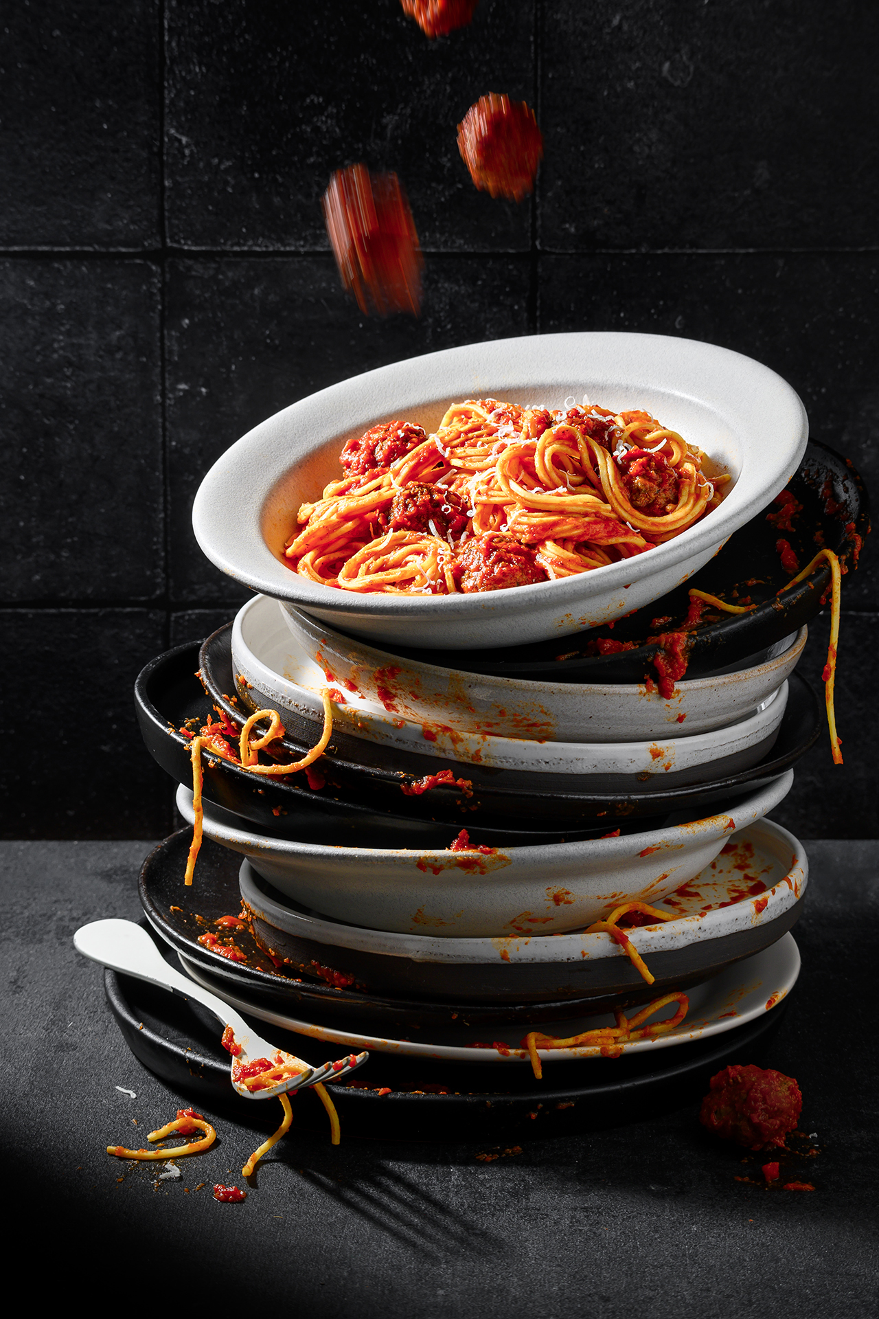 A chaotic stack of dirty plates, topped with spaghetti and meatballs, with pasta falling off, set against a dark background