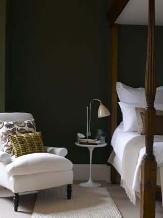 grey and white bedroom with dark grey walls, four poster bed, white armchair, patterned cushions, side table, lamp