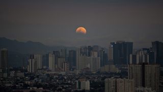 A reddish moon is pictured rising above a city. The lower part of its disk is hidden by Earth's shadow during an eclipse.