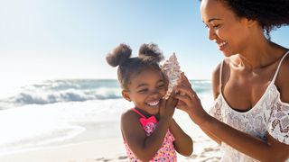 A girl and her mother are at the beach. The mother holds a seashell to her daughter's ear.