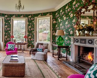 A classic living room with green patterned wallpaper, fuchsia pink chair and brass framed mirror above mantelpiece and fireplace