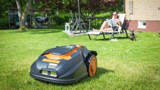 A robot lawn mower mowing the grass with someone sitting in a lawn chair in the background