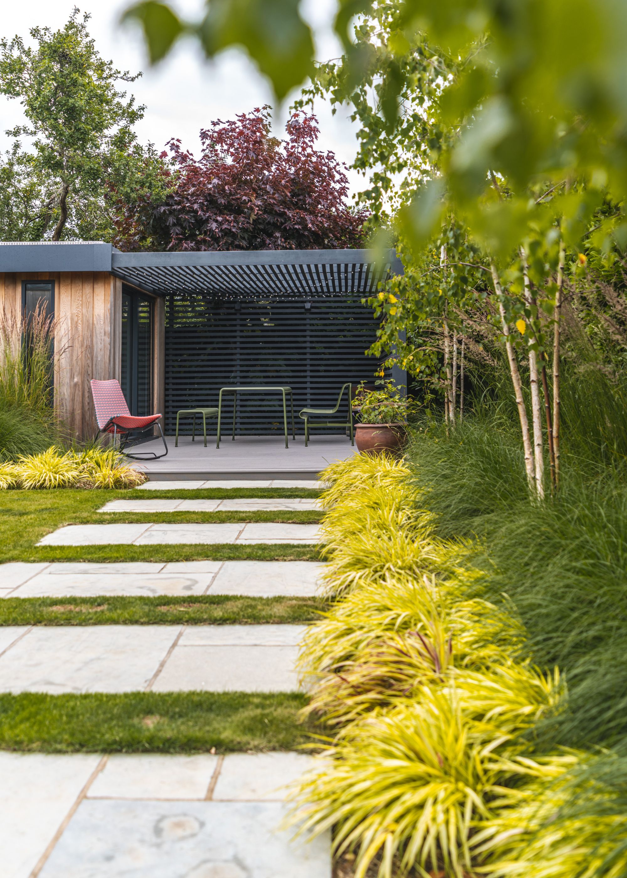 A lush green garden with lots of plants by the right side of the fence. There is a paved pathway with lots of plants between and a view of outdoor patio furniture.