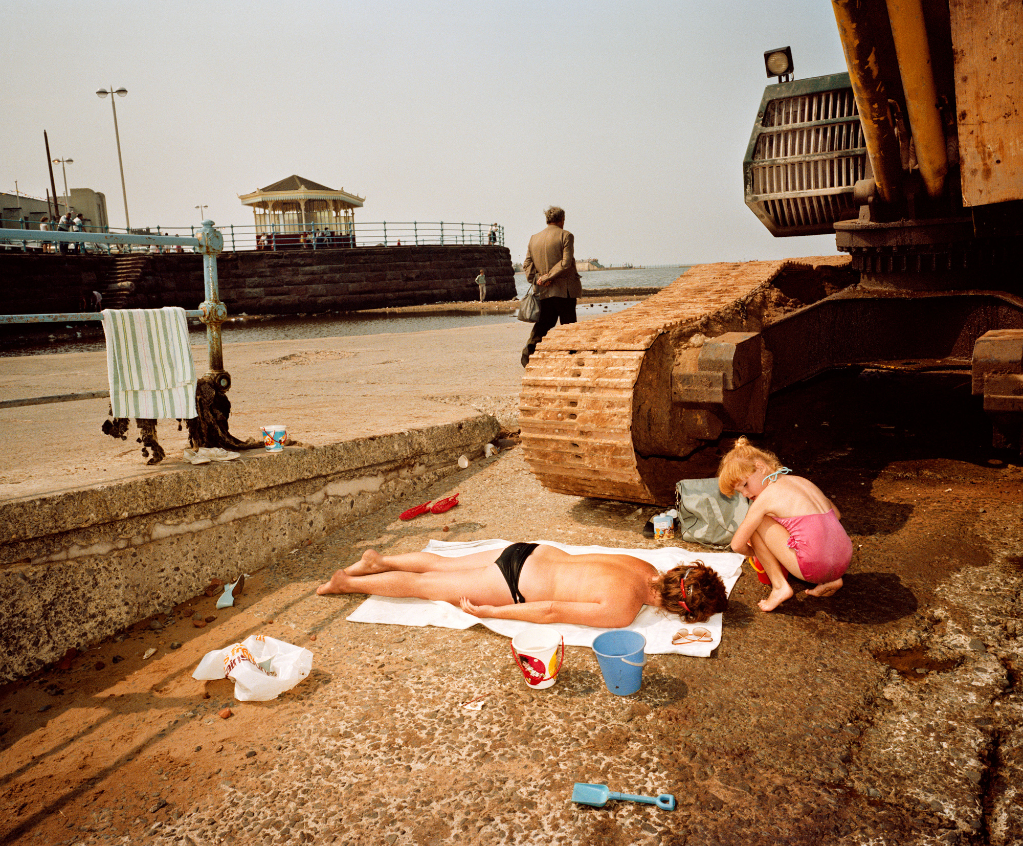 A young child kneels on the ground next to a woman lying on a towel in the sun next to a large piece of rusty machinery on a paved area near a body of water.