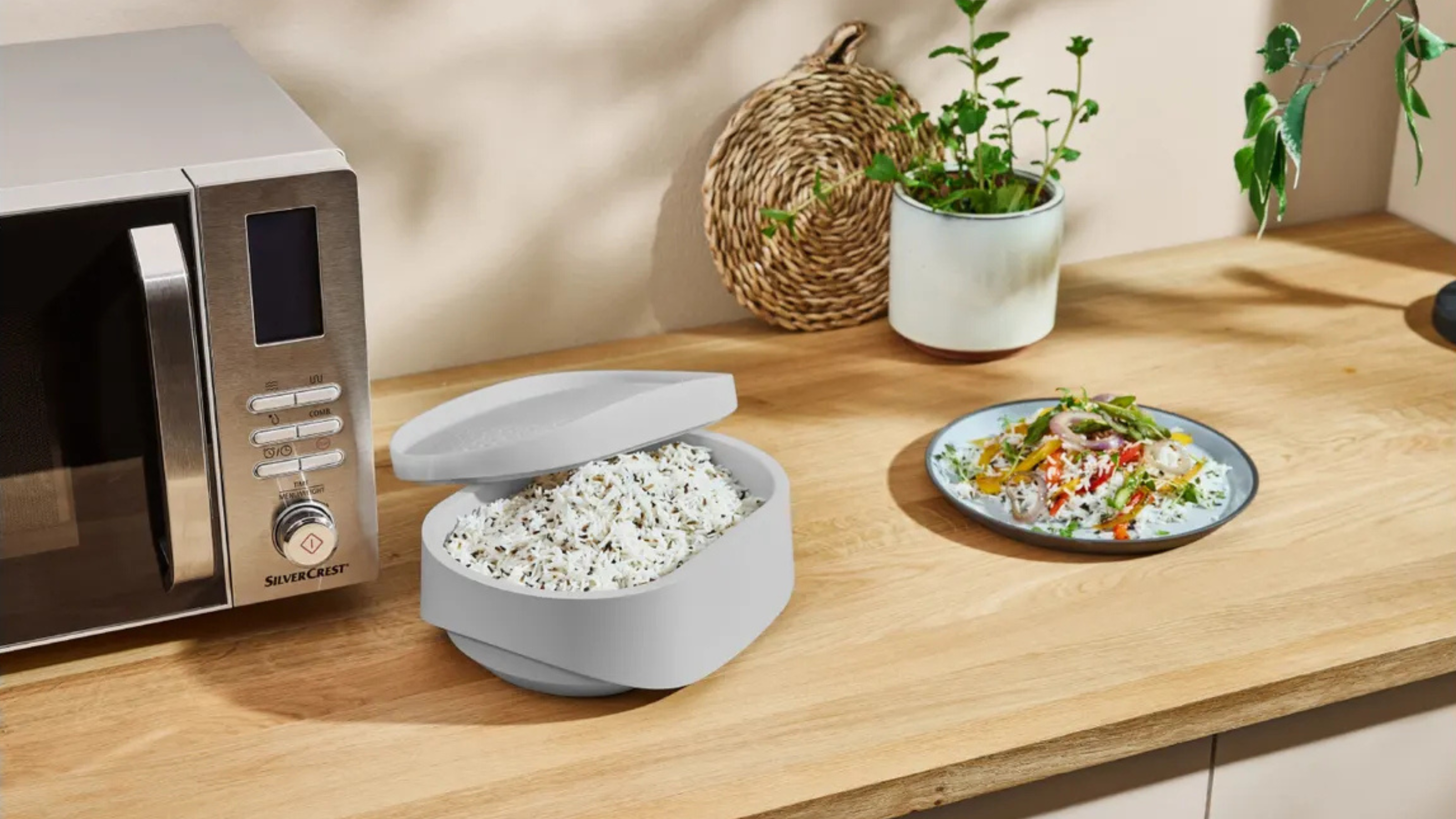 The Silvercrest Microwave Rice Cooker next to microwave and cooked plate on rice on a brown, wooden kitchen worktop.