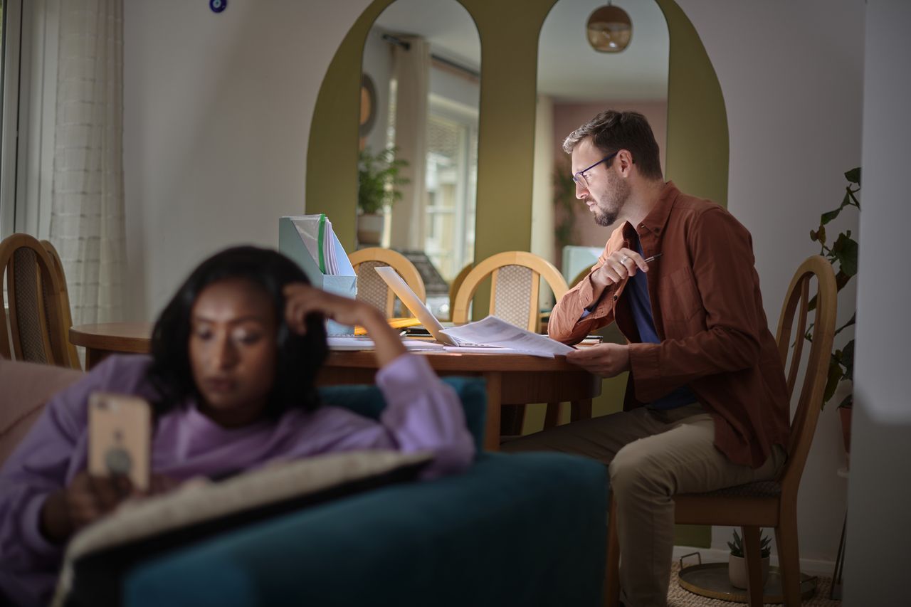 A young man looks at personal finance papers at a kitchen table while a young woman looks at her phone on the couch.