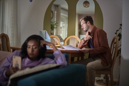 A young man looks at personal finance papers at a kitchen table while a young woman looks at her phone on the couch.