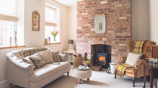 a cosy country living room with two windows, a grey sofa with beige and green pillows, a brick fireplace and an armchair with a tartan blanket