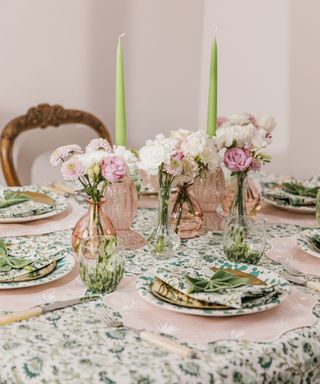 A green and white floral patterned table with green plates, pink wavy table mats, and clear and pink vases of flowers and green candlesticks