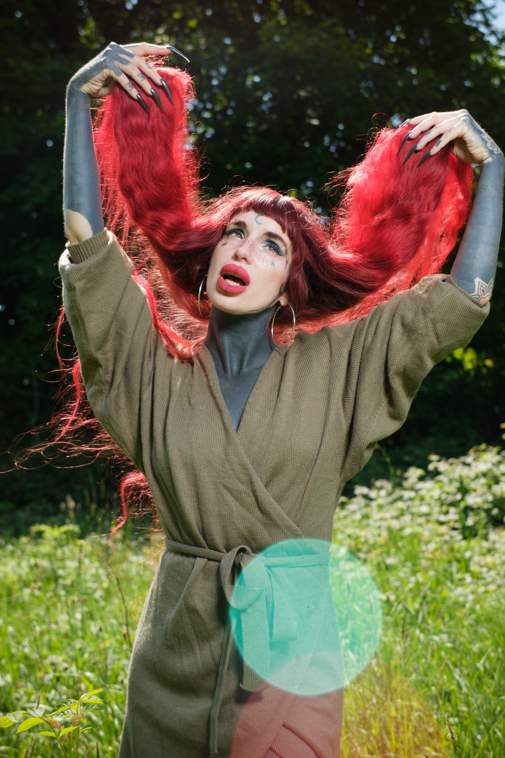 Red-haired person with dark body paint holding up their hair in bright sunlight, with a large turquoise lens flare across the lower frame.