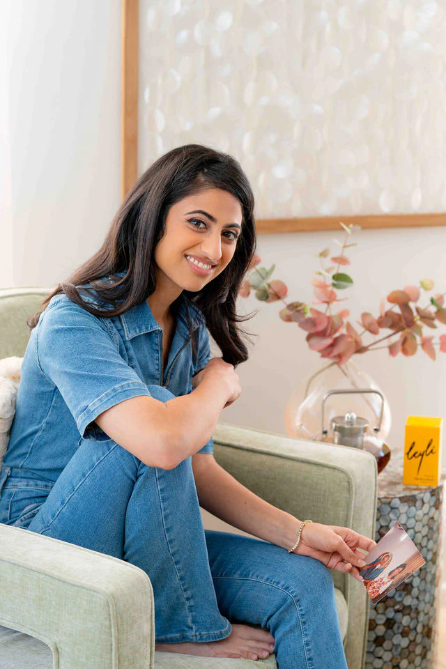 A woman of color with dark brown hair pictured sitting on a pale green accent chair, holding a picture of herself as a child with her Persian grandmother