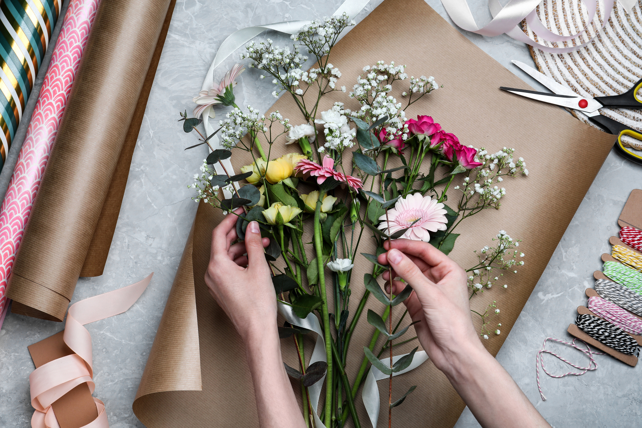 Top view of a woman arranging flowers with brown paper, ribbon, and string.