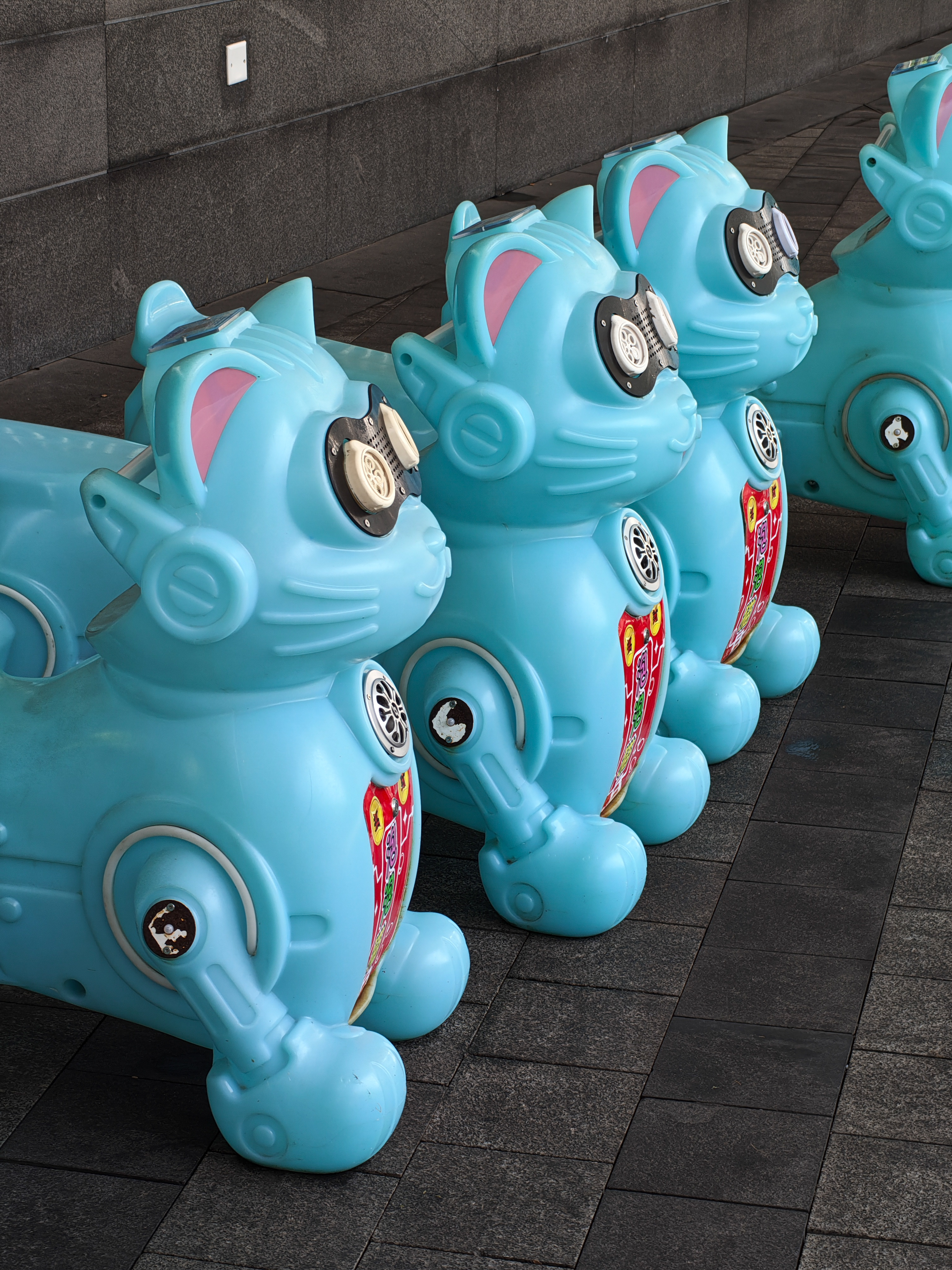 A row of identical, bright blue plastic toy rides shaped like stylized robotic cats. They are lined up on a dark gray tiled walkway. Each "cat" features pink ears, black-rimmed "eyes" that look like speakers, and a red decorative panel on its chest. The perspective shows the toys receding into the distance against a dark stone wall.