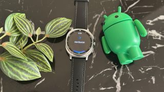 The Gemini app on the Samsung Galaxy Watch 8 Classic, which lays on a black marble table next to plant leaves and an Android mascot figurine.