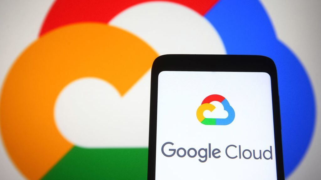 A phone, bearing the Google Cloud logo and the words &amp;#039;Google Cloud&amp;#039;, is held in front of a wall bearing a larger version of the same logo