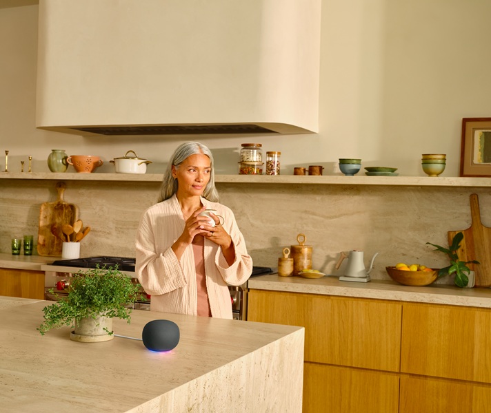 The new Google Home Speaker in black, sitting on a counter top with a model standing beside it, holding a mug.