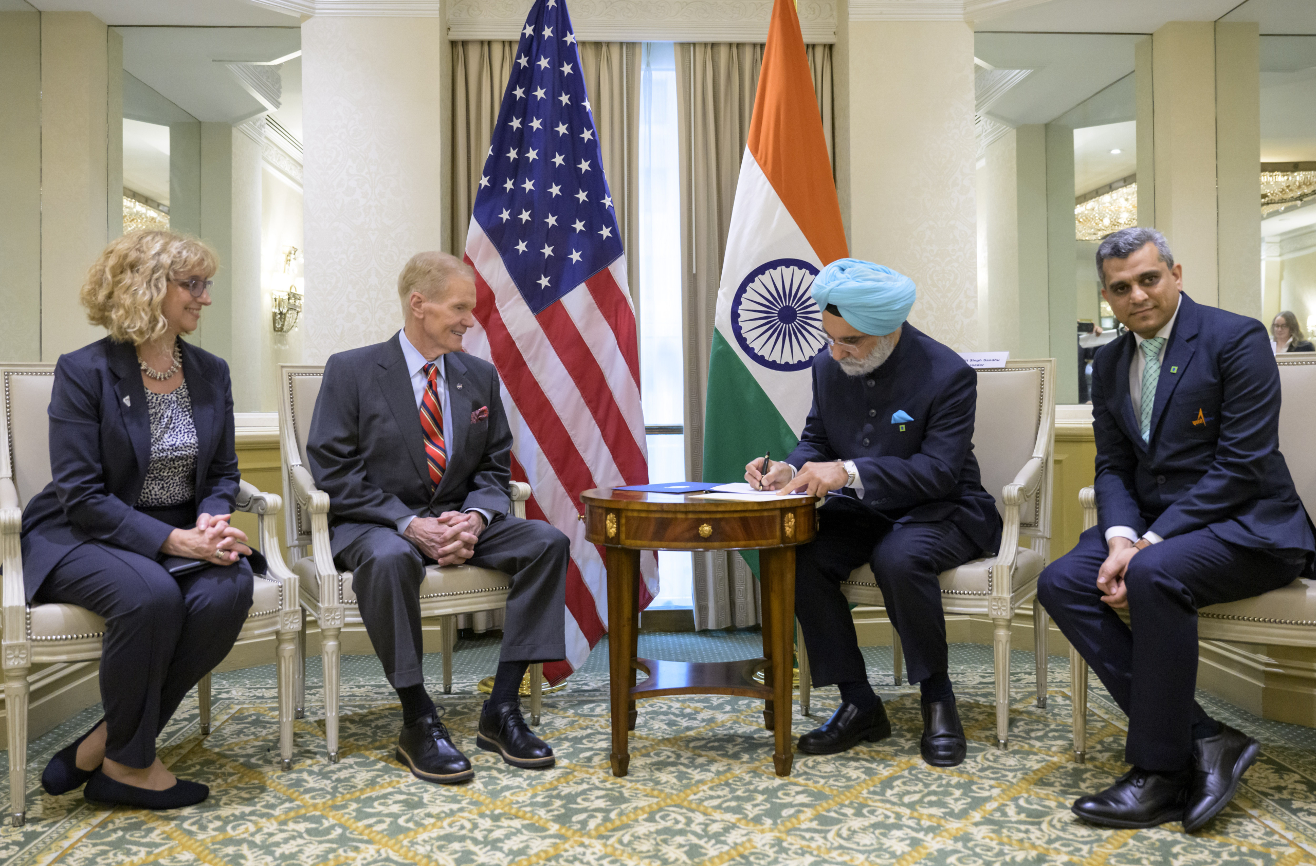 Four people sit facing forward, not facing each other.  They all wear suits.  A man in a light blue turban signs a document on a small table in front of him.  Two flags stand behind two people in the middle.