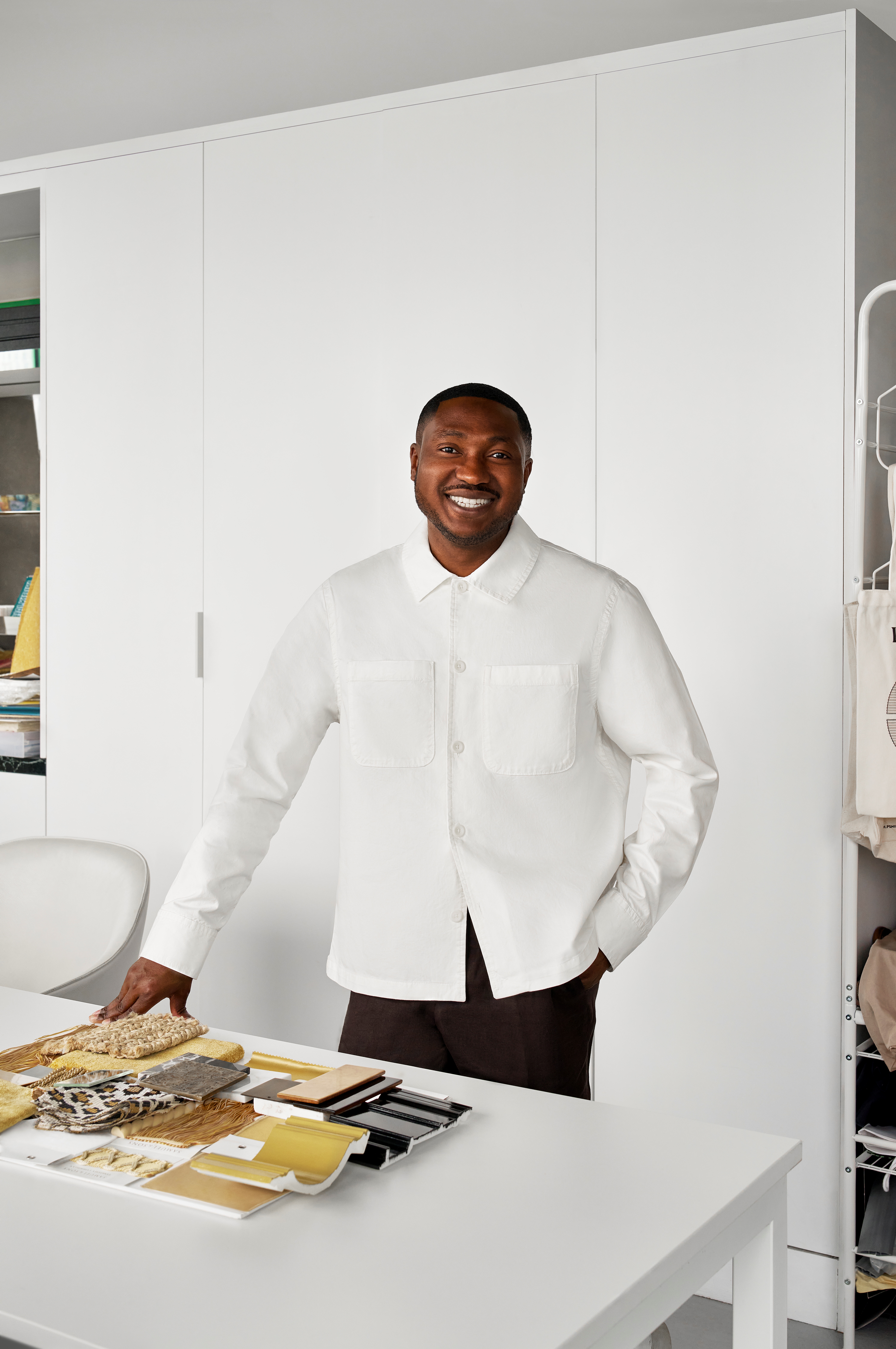 A young man dressed in a white shirt and black trousers stands in a design showroom with all neutrally tinted furnishings in front of a series of material samples while smiling at the camera.